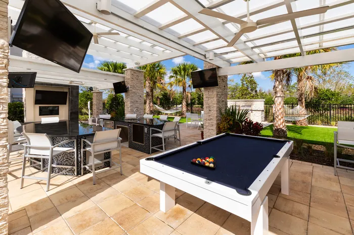 Luxurious outdoor living space featuring a sleek pool table, granite bar, and modern seating area under a pergola with lush landscaping.