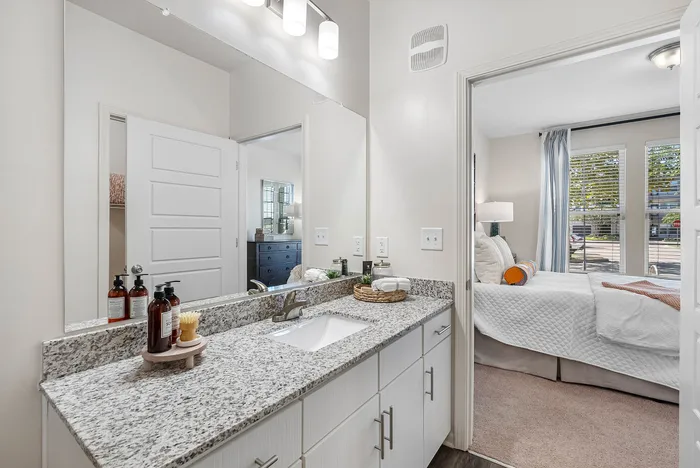 Bright, modern bathroom with granite countertop leads to a cozy bedroom featuring large windows and stylish decor.
