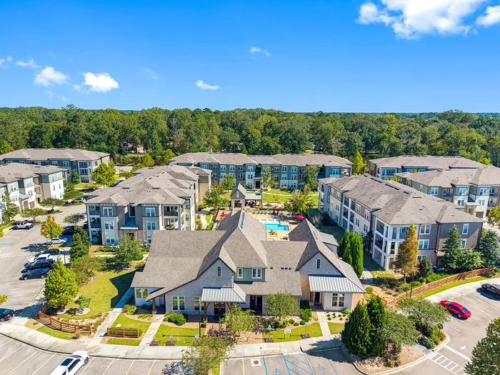 Aerial view of a charming apartment complex surrounded by lush greenery, featuring modern buildings, ample parking, and a central pool area under a clear blue sky.