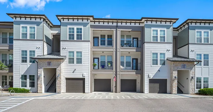 Modern apartment building with three stories and balconies, featuring a stylish stone entryway and integrated garages, set under a vibrant blue sky.