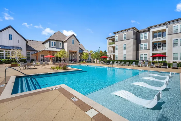 Luxurious apartment complex featuring a sparkling pool with modern loungers and elegant surrounding architecture under a clear blue sky.