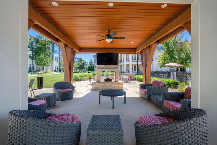 Charming outdoor lounge area with stylish wicker seating under a wooden pergola, featuring a modern ceiling fan and an inviting fireplace, perfect for relaxation and social gatherings.