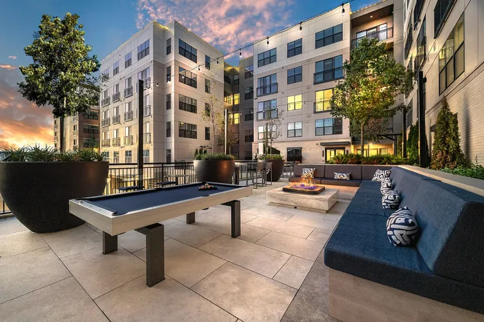 Luxurious apartment courtyard featuring a modern pool table, stylish seating with cushions, ambient string lighting, and lush greenery under a stunning sunset sky.