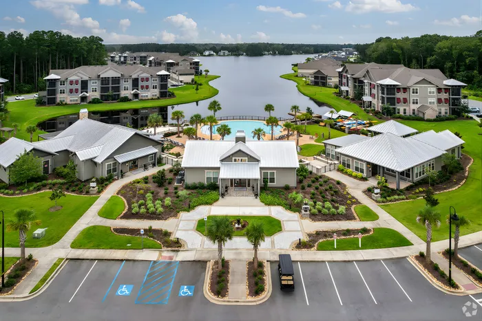 Aerial view of luxury lakeside apartment community featuring modern buildings, lush green landscaping, and a central pool area surrounded by palm trees.