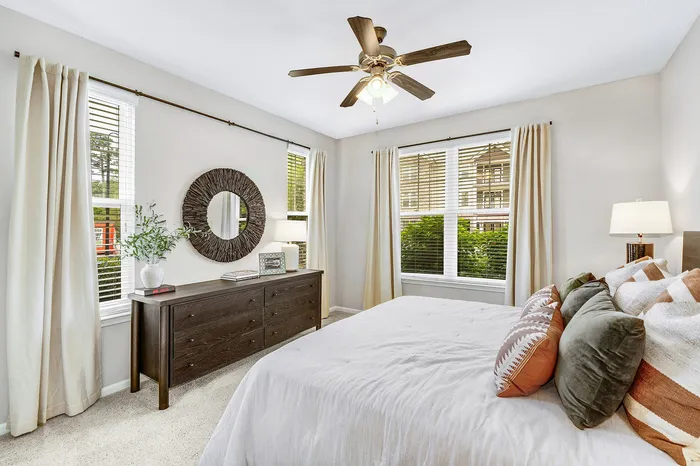 Bright and inviting bedroom featuring a chic ceiling fan, large windows with serene views, plush bedding, and elegant decor.