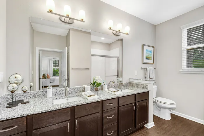 Elegant bathroom with double vanity, granite countertops, and modern lighting, leading into a bright bedroom.