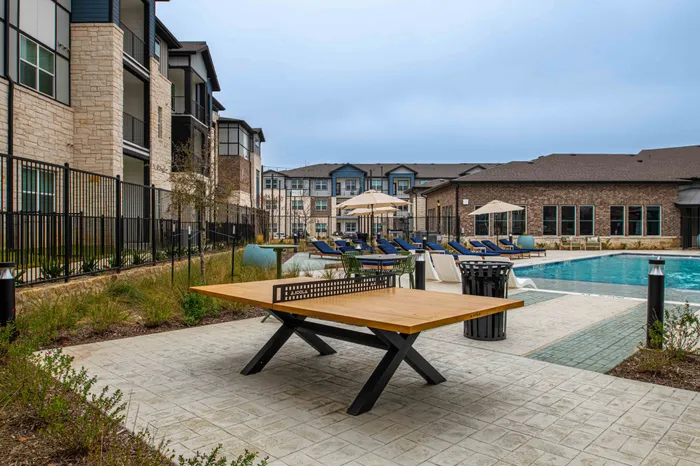 Modern apartment complex exterior featuring elegant architecture, a sparkling swimming pool with lounge chairs, and a stylish outdoor ping pong table.