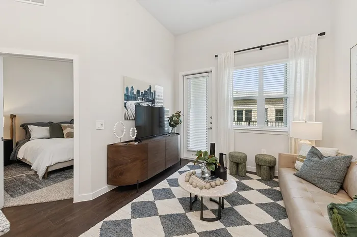 Charming living room with modern decor, featuring a cozy beige couch, stylish rug, and natural light through large windows. Includes a glimpse of a well-appointed bedroom.