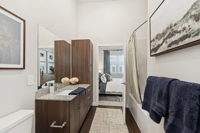 Elegant bathroom with modern fixtures, granite countertop, and stylish wood cabinetry, leading to a cozy bedroom with plush bedding and natural light.