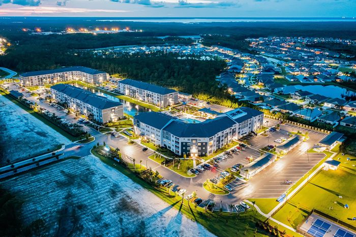 Stunning aerial view of a modern apartment complex illuminated at dusk, showcasing vibrant community spaces, ample parking, and beautiful landscaping in a thriving neighborhood.