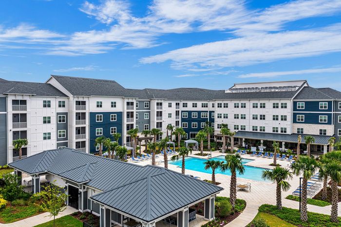 Luxurious apartment complex with a modern exterior, pristine swimming pool, and lush palm trees, set against a bright blue sky.