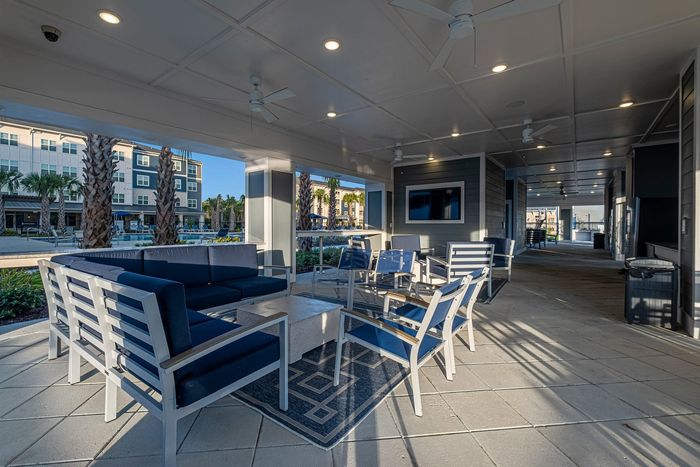 Elegant covered outdoor seating area with modern lounge furniture, ceiling fans, and scenic views of palm trees and a stylish apartment complex.