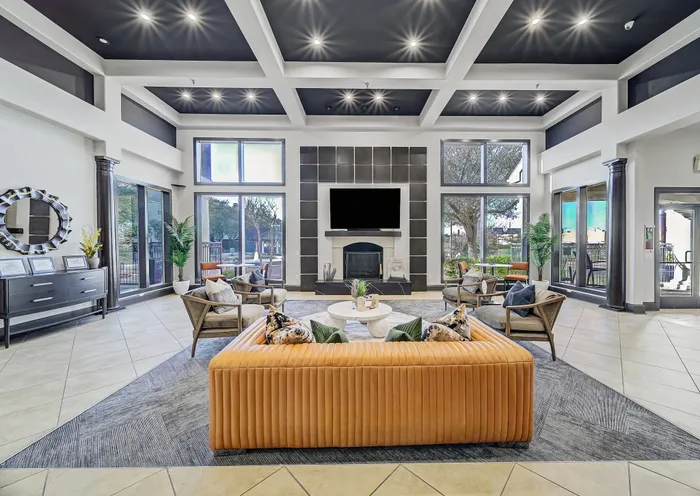Elegant apartment clubhouse with modern decor, featuring plush seating around a central fireplace, high ceilings, and large windows offering natural light.