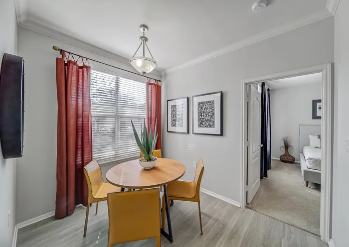 Bright dining area with a modern round wooden table, stylish yellow chairs, and large windows with red curtains. Tastefully decorated with framed artwork and a plant centerpiece.