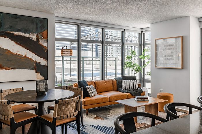 Modern living area with a stylish brown leather sofa, large abstract artwork, and floor-to-ceiling windows offering city views.