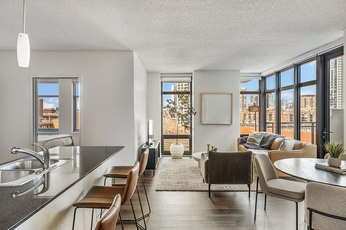 Bright modern apartment living space with a sleek kitchen island, open seating area, and floor-to-ceiling windows offering stunning cityscape views.