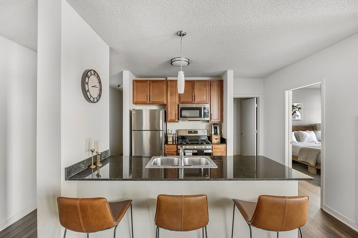 Modern kitchen with chic bar seating, stainless steel appliances, and warm wood cabinetry; adjacent cozy bedroom visible.