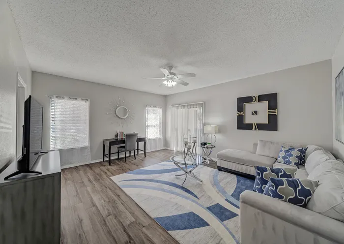 Spacious and modern living room with elegant white sofa, stylish geometric rug, and sleek glass coffee table, bathed in natural light from large windows in a contemporary apartment.