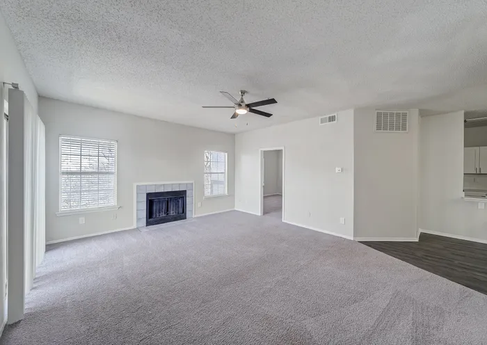 Spacious living room with plush carpeting, cozy fireplace, and modern ceiling fan, leading into an inviting bedroom and kitchen area.
