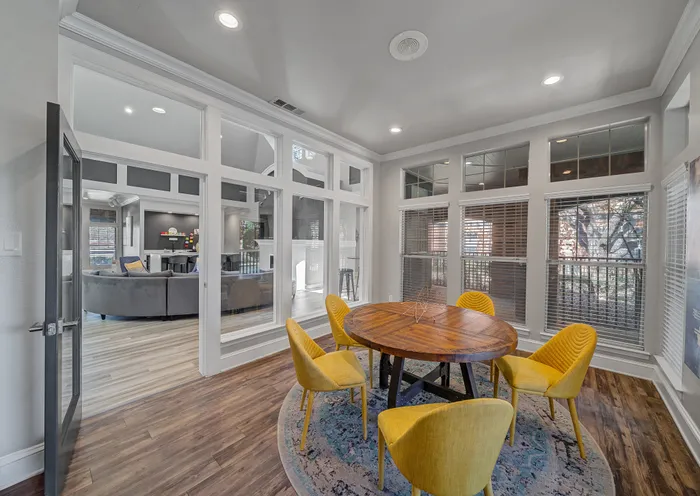 Bright and inviting dining area with a round wooden table and vibrant yellow chairs, featuring large windows providing ample natural light and a view into a stylish living space.