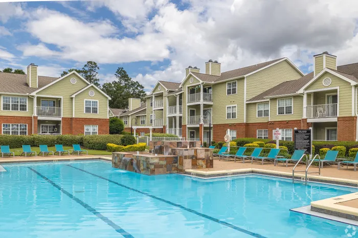 Inviting apartment complex with modern architecture, large swimming pool, and blue lounge chairs under a vibrant blue sky.