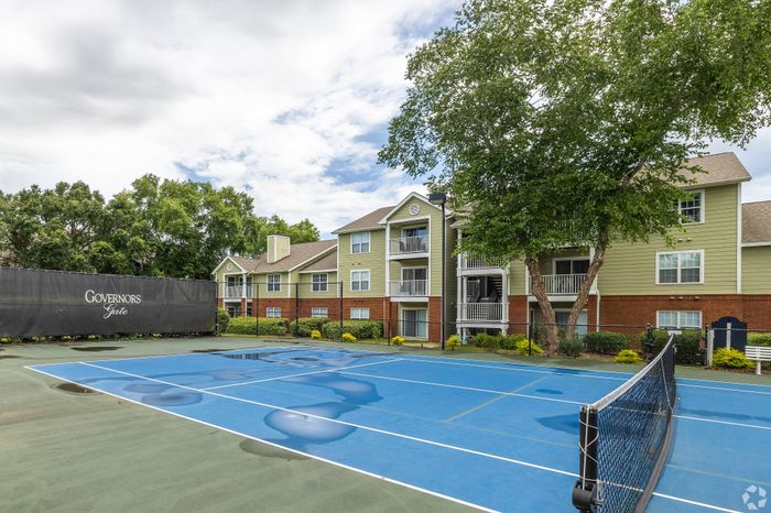 Inviting apartment complex featuring vibrant green buildings with balconies, nestled next to a well-maintained tennis court and surrounded by lush trees.