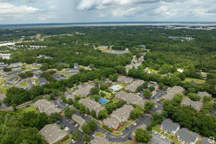 Vibrant aerial view of a lush residential community nestled among green trees, featuring multiple modern apartment buildings, a sparkling swimming pool, and tennis courts, offering a serene suburban lifestyle.