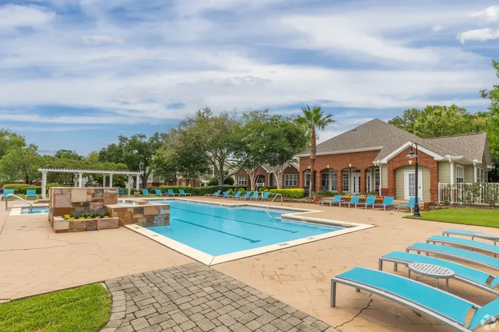 Inviting outdoor pool area featuring modern lounge chairs, surrounded by lush greenery and a charming brick clubhouse.