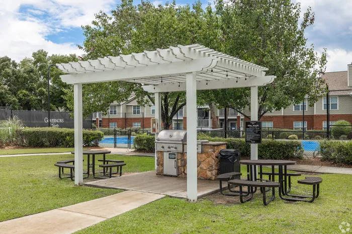 Charming outdoor picnic area with a stylish pergola, modern grill, and inviting seating. Perfect for enjoying sunny days by the pool in a lush, green setting.