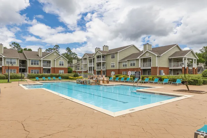 Luxurious apartment complex featuring a sparkling blue swimming pool surrounded by comfortable lounge chairs, vibrant greenery, and elegant multi-story buildings under a picturesque sky.