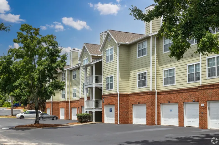 Charming multi-story apartment complex with attached garages, surrounded by lush greenery and a bright blue sky.