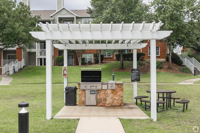 Charming outdoor barbecue area with a modern grill under a white pergola, set against the backdrop of a welcoming apartment complex and lush greenery.