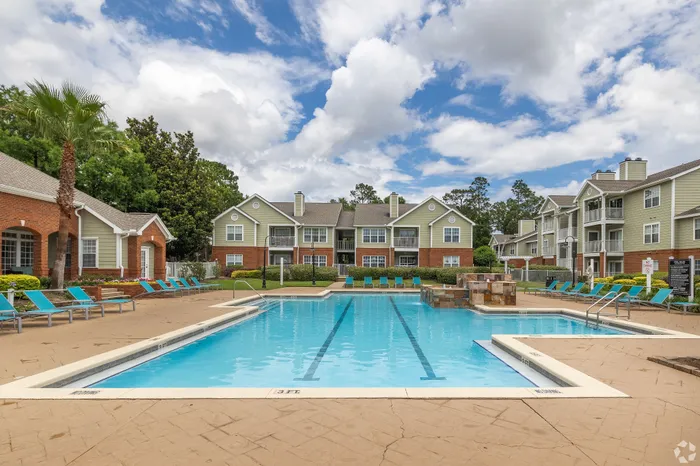 Charming apartment complex with a pristine swimming pool, surrounded by lush greenery and stylish lounge chairs under a bright blue sky.