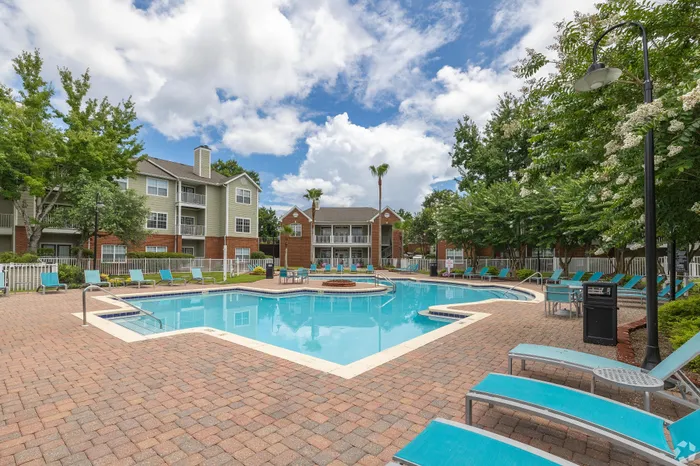 Vibrant apartment community showcasing a sparkling swimming pool, surrounded by lush greenery and inviting lounge chairs, against a backdrop of modern residential buildings with bright skies.
