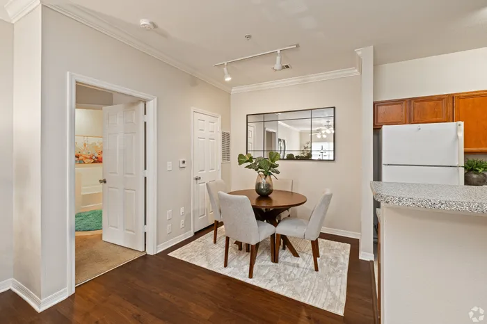 Cozy dining area with a round table and four soft chairs, complemented by a decorative vase and a large mirror reflecting natural light. Warm wooden floors enhance the inviting atmosphere.