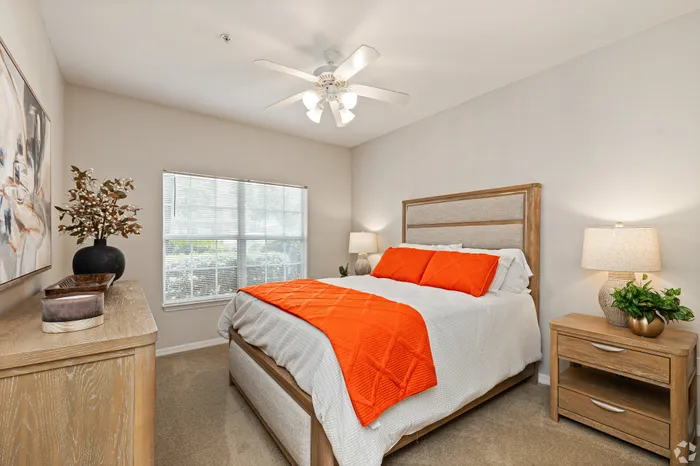 Bright and inviting bedroom featuring a cozy bed with vibrant orange accents, modern ceiling fan, and stylish wooden furniture.