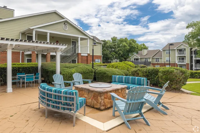 Charming apartment complex featuring stylish outdoor seating around a cozy fire pit, with manicured gardens and elegant architecture under a bright blue sky.