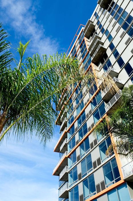 Modern high-rise building with sleek balconies and large glass windows, complemented by lush palm trees and a vibrant blue sky backdrop.