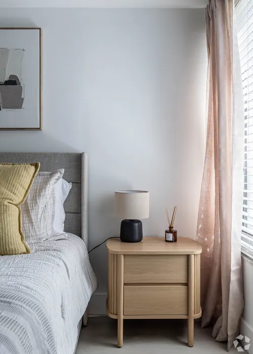 Elegant bedroom corner featuring a cozy bed with plush pillows, a stylish wooden nightstand, and a soft-curtain-draped window inviting natural light.