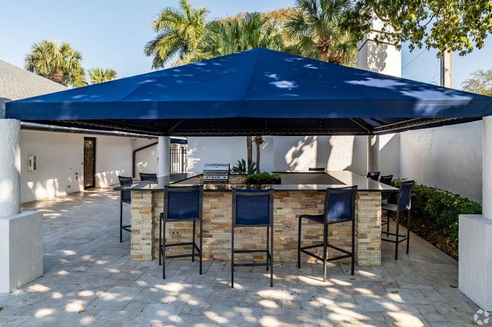 Charming outdoor patio area with a stylish stone bar, shaded by a blue canopy, perfect for entertaining guests amidst lush greenery and palm trees.