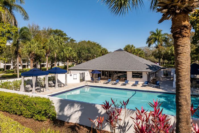 Luxurious apartment amenities featuring a sparkling pool, elegant lounge chairs, and shaded cabanas set amidst lush palm trees.