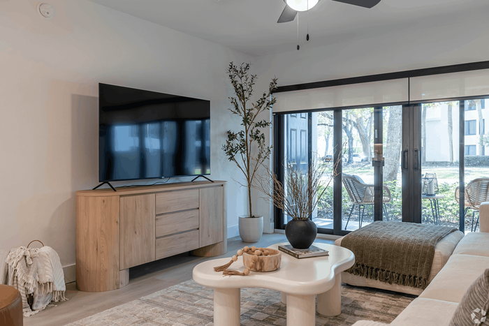 Chic living room with modern decor, large flat-screen TV on a wooden console, cozy seating, and floor-to-ceiling windows opening to a serene outdoor view.