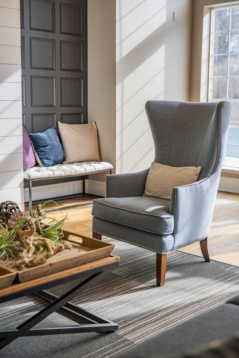 Bright and inviting living space featuring a cozy gray wingback chair, stylish pillows on a bench, and a modern coffee table with decorative plants.