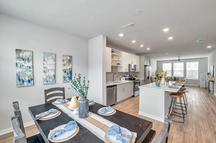 Bright and modern open-concept dining and kitchen area with sleek cabinetry, stainless steel appliances, and elegant decor, leading into a spacious living room with large windows.