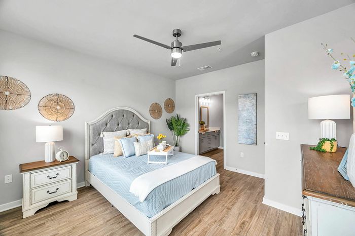 Inviting bedroom with plush bedding, light wood flooring, modern ceiling fan, and elegant decor, seamlessly connecting to a chic bathroom.