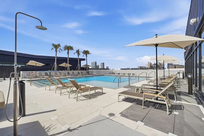 Luxurious rooftop pool area with sleek sun loungers and large umbrellas, offering a stunning city skyline view and a perfect relaxation spot under the clear blue sky.