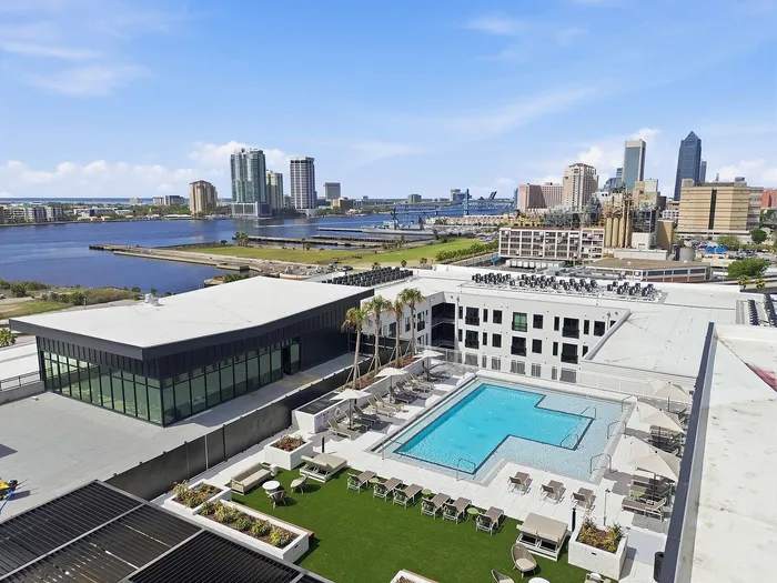 Stunning aerial view of a luxurious apartment complex with a sparkling pool, modern amenities, and a scenic waterfront city skyline.