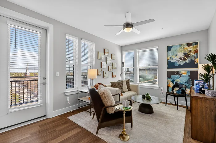 Chic, sunlit living room with stylish decor, featuring floor-to-ceiling windows, artful wall displays, and a cozy seating area with modern furnishings.