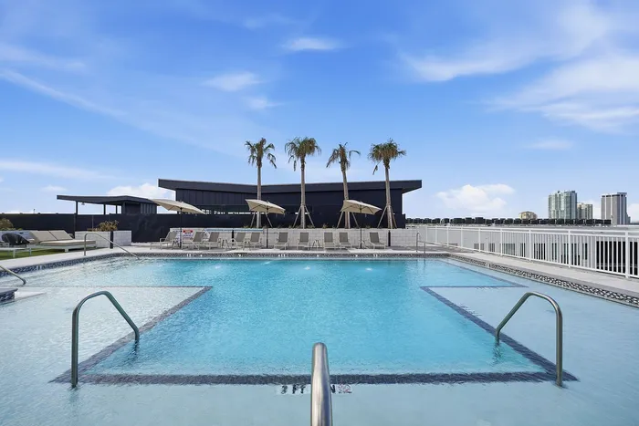 Luxurious rooftop swimming pool with palm trees and sun loungers, offering stunning city views and a serene blue sky.