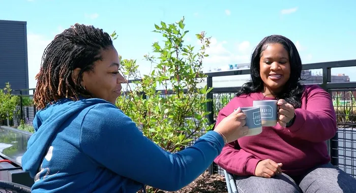 Two people enjoying coffee on a sunny apartment balcony, surrounded by lush greenery and city views.